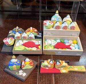 手前左/ひな人形(一対6500円、杉板の台座付き)。手前右/犬筥(小、一対8500円、羽子板付き)。奥/ひな人形、三人官女、陶菓子、福良雀(ふくらすずめ)のセット(1万9500円〜、桐箱付きもあり)